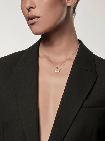 A person wearing a deep V-neck black blazer with a minimalist design is adorned with two delicate necklaces. One necklace features the Three Letters Necklace in 18k Gold, showcasing small circular pendants and charms. The background is plain white, emphasizing the sophisticated look.