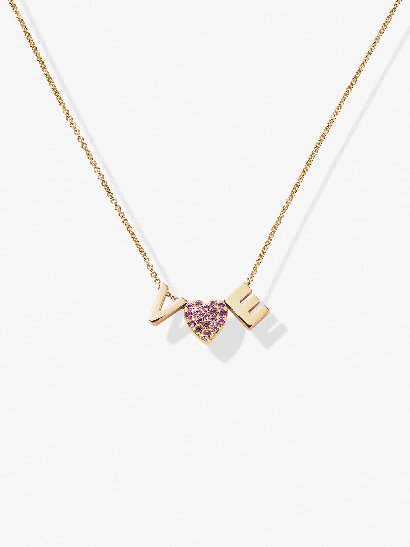 A delicate Two Letters and Pink Sapphire Heart Necklace in 18k Gold features three hanging pendants with two gold letters "VE." and "heart "the heart is set with vibrant hued pink sapphires, encrusted in gemstones. The necklace lies gracefully on a white background.