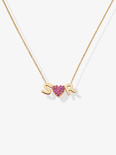 The Two Letters and Ruby Heart Necklace in 18k Gold is a delicate piece, showcasing letter charms "S" and "R" flanking a small ruby heart adorned with pink gemstones. The heart is elegantly positioned between the two letters, all displayed against a plain white background.