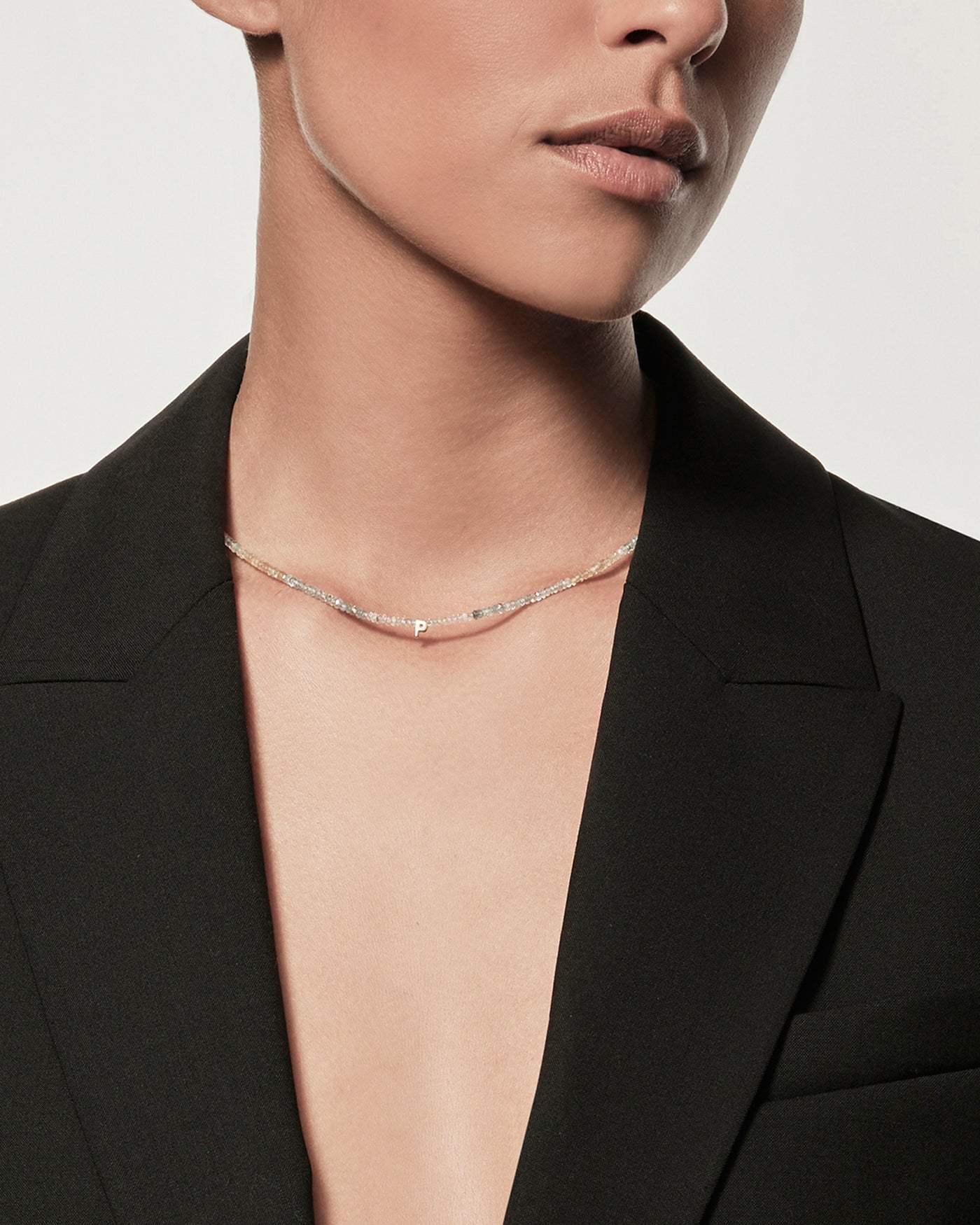 A person with medium skin tone is wearing an open black blazer without a shirt underneath and a delicate One Letter Necklace in Aquamarine gemstones and 9k Gold. The background is plain and out of focus, highlighting the attire and accessories. The person's face is partly visible, focusing on their lower half.