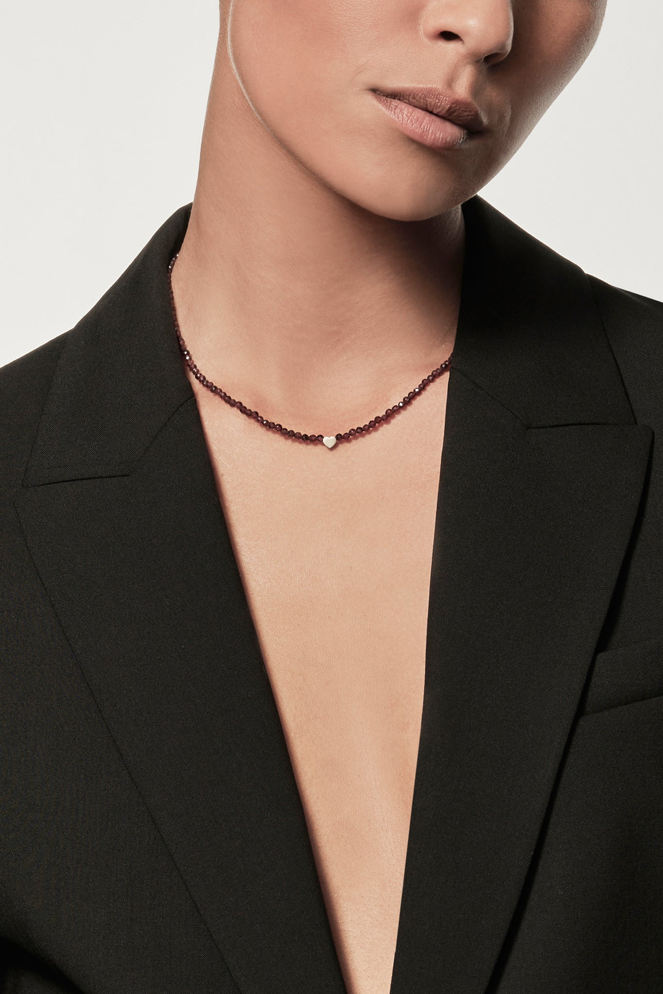 A person is shown from the neck down, donning a deep V-neck blazer over a bare chest. They're accessorised with the Heart Necklace in Garnet and 9k Gold, adding an elegant touch to their ensemble. The plain light background accentuates the minimalistic and sophisticated fashion style.
