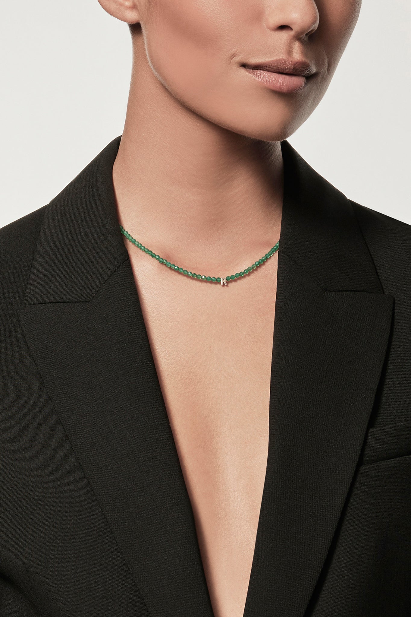 Cropped image of a person wearing a black blazer with no shirt underneath, showcasing the One Letter Necklace in Green Onyx and 9k Gold. The background is plain white.