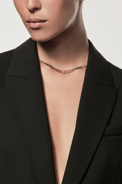 A person with medium skin tone is wearing an open black blazer without a shirt underneath and a delicate One Letter Necklace in Tourmaline and 9k Gold. The background is plain and out of focus, highlighting the attire and accessories. The person's face is partly visible, focusing on their lower half.