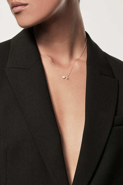 A person wearing an open black blazer and a delicate One Heart and Letter Necklace in Diamonds and 18k Gold, posing against a plain background. Their head is slightly turned to the side, with short hair styled neatly. The image emphasises style and elegance.
