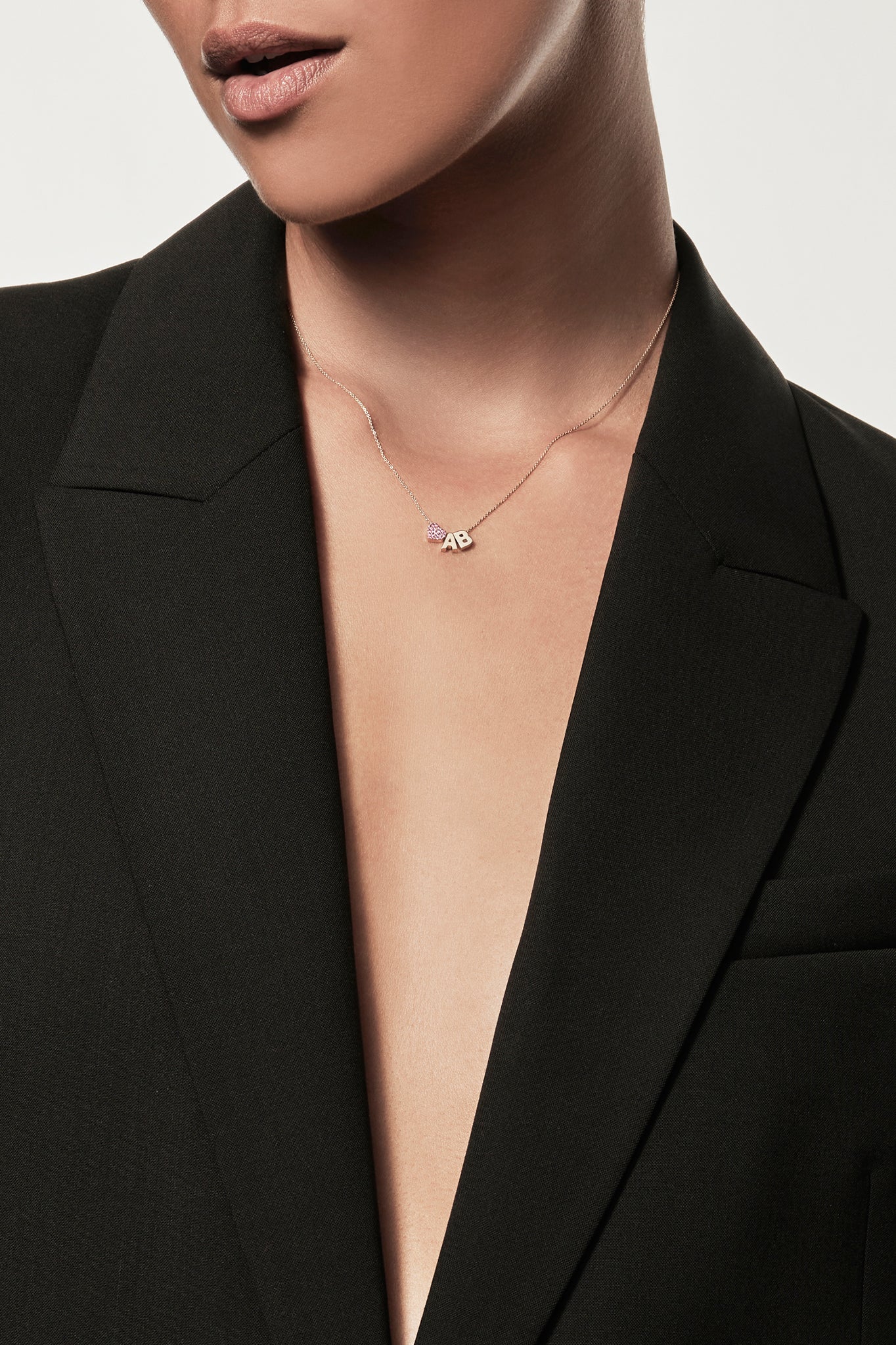 Close-up of a woman wearing a minimalist gold initial “A” and "B" separated with a pink sapphire heart charm, styled with a black blazer – personalised alphabet pendant jewellery