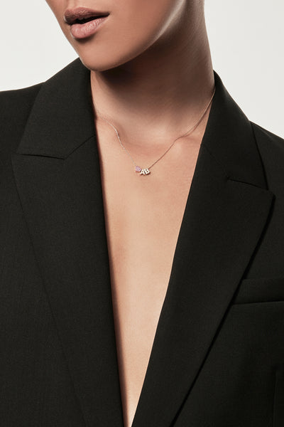 Close-up of a woman wearing a minimalist gold initial “A” and "B" separated with a pink sapphire heart charm, styled with a black blazer – personalised alphabet pendant jewellery