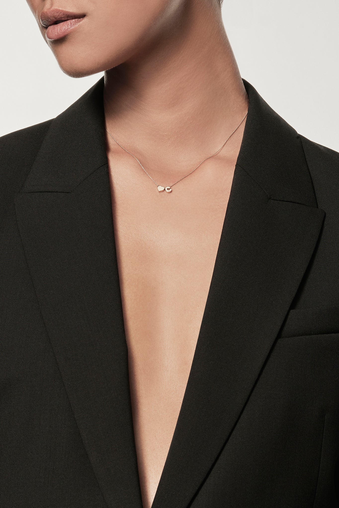 Close-up of a woman wearing a minimalist 18k gold initial "C" and gold heart charm  on a fine chain, styled with a black blazer – personalised alphabet pendant jewellery