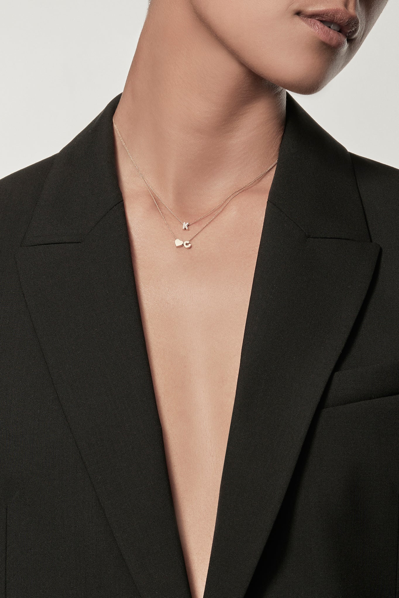 Close-up of a woman wearing a minimalist 18k gold initial "C" and gold heart charm  on a fine chain, layered with a diamond letter "K" necklace, styled with a black blazer – personalised alphabet pendant jewellery