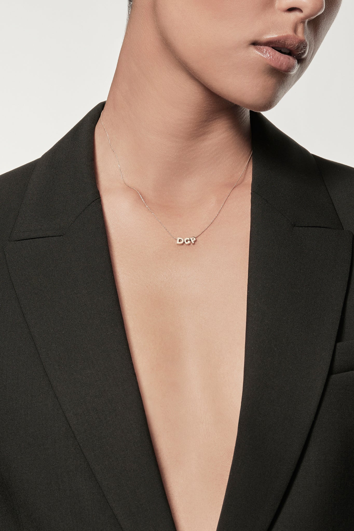 Close-up of a woman wearing a minimalist 18k gold initials “D" "C" "P" necklace and diamond "G" necklace on a fine chain, styled with a black blazer – personalised alphabet pendant jewellery