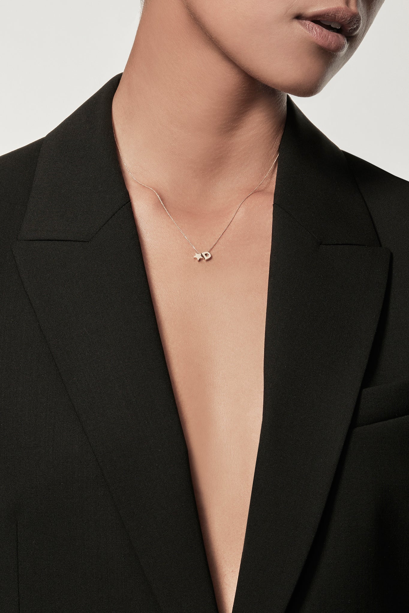 Close-up of a woman wearing a minimalist 18k gold initial “C” and gold star necklace on a fine chain, styled with a black blazer – personalised alphabet pendant jewellery