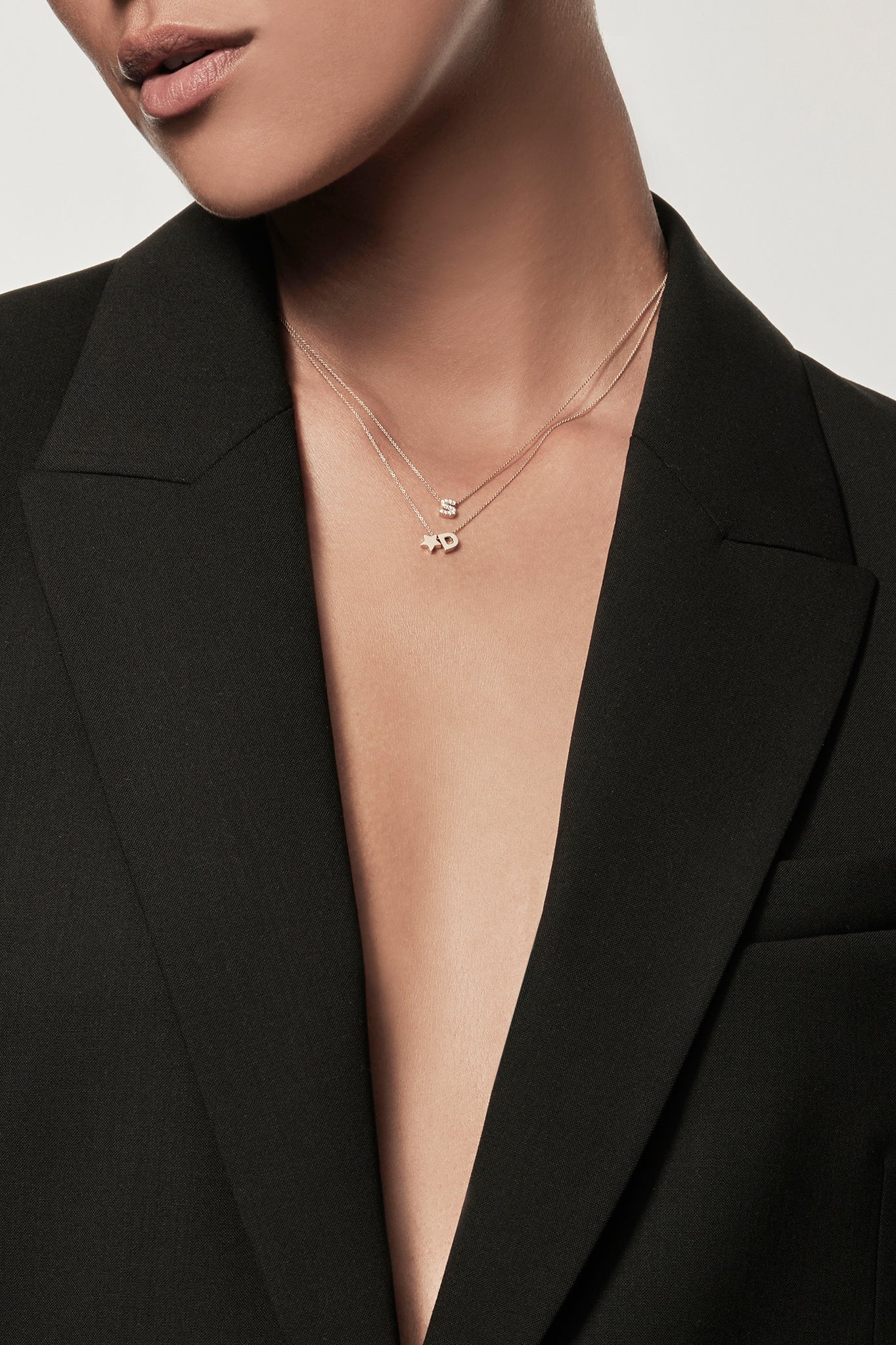 Close-up of a woman wearing a minimalist 18k diamond initial “S”  and gold star charm with initial "D" layered necklaces on fine chains, styled with a black blazer – personalised alphabet pendant jewellery