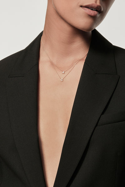 Close-up of a woman wearing a minimalist 18k gold initial “A” necklace and diamond initial "C" necklace on a fine chain, styled with a black blazer – personalised alphabet pendant jewellery