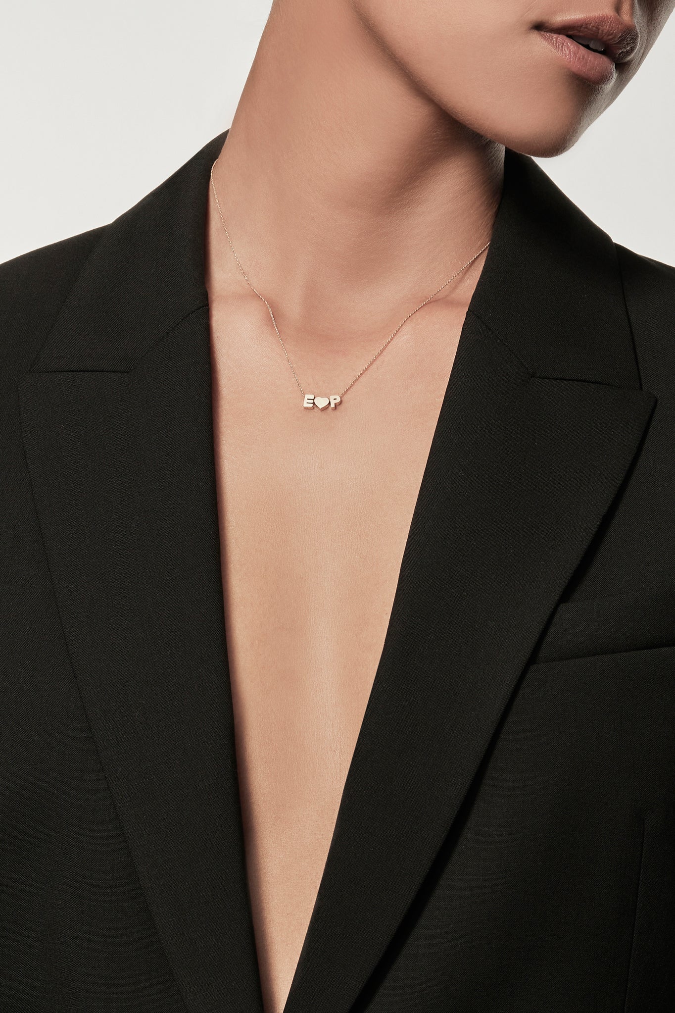 Close-up of a woman wearing a minimalist 18k gold initial “A” gold heart charm gold initial "P"  on a gold chain, styled with a black blazer – personalised alphabet pendant jewellery