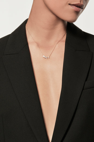 A person wearing an open black blazer and a delicate One Heart and Letter Necklace in Diamonds and 18k Gold, posing against a plain background. Their head is slightly turned to the side, with short hair styled neatly. The image emphasizes style and elegance.
