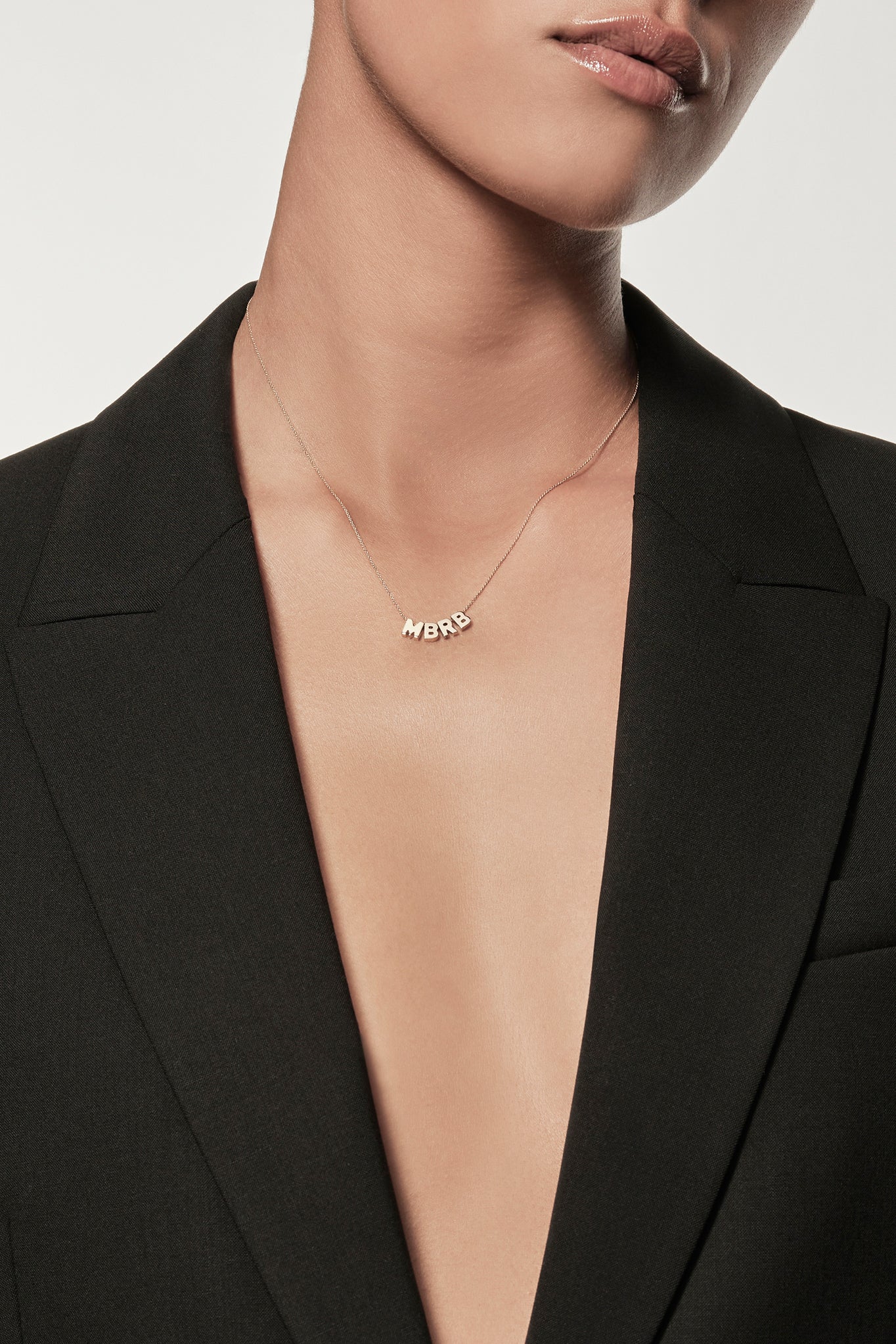 Close-up of a woman wearing a minimalist 18k gold initials “M” "B" "R" "B" necklace and diamond love spinner pendant necklace on a fine chain, styled with a black blazer – personalised alphabet pendant jewellery
