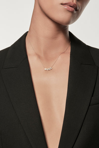 Close-up of a woman wearing a minimalist 18k gold initials “M” "B" "R" "B" necklace and diamond love spinner pendant necklace on a fine chain, styled with a black blazer – personalised alphabet pendant jewellery
