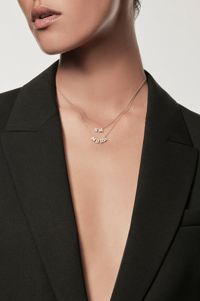 Close-up of a woman wearing a minimalist 18k gold initials “M” "B" "R" "B" necklace and diamond initials "S" "M" necklace on a fine chain, layered with a black blazer – personalised alphabet pendant jewellery