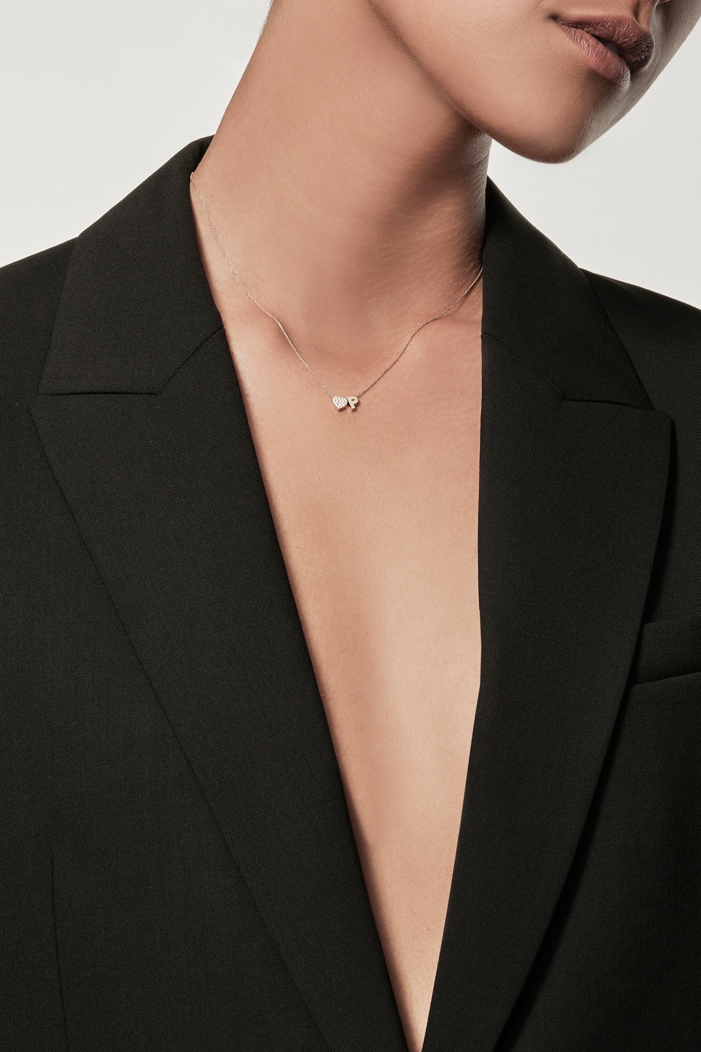 Close-up of a woman wearing a minimalist 18k gold and lab-grown diamond heart charm and gold "P" letter charm on a fine chain, styled with a black blazer – personalised alphabet pendant jewellery