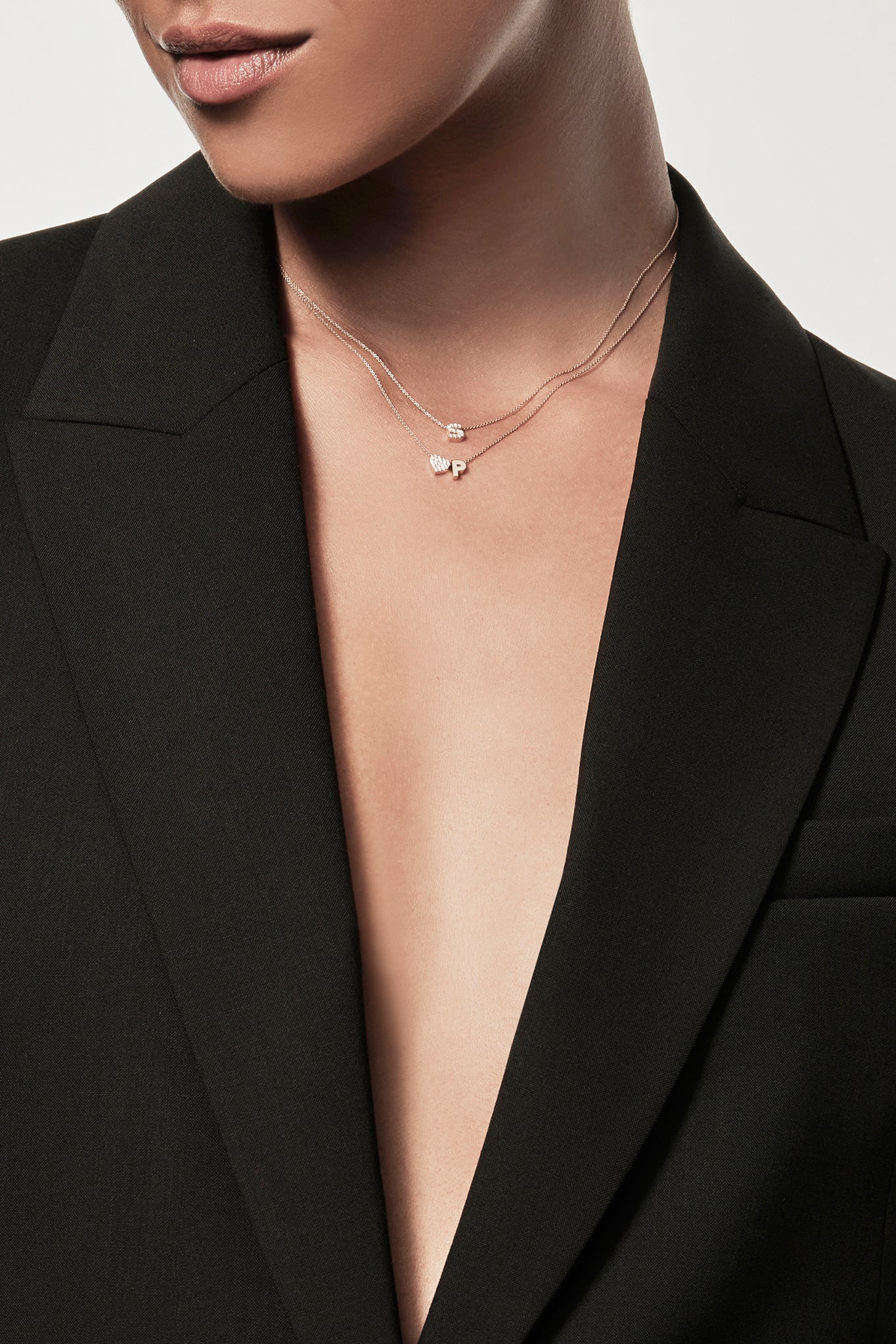 Close-up of a woman wearing a minimalist 18k gold and lab-grown diamond initial "S"  and lab-grown diamond heart and gold "P" letter charm on a fine chain, styled with a black blazer – personalised alphabet pendant jewellery
