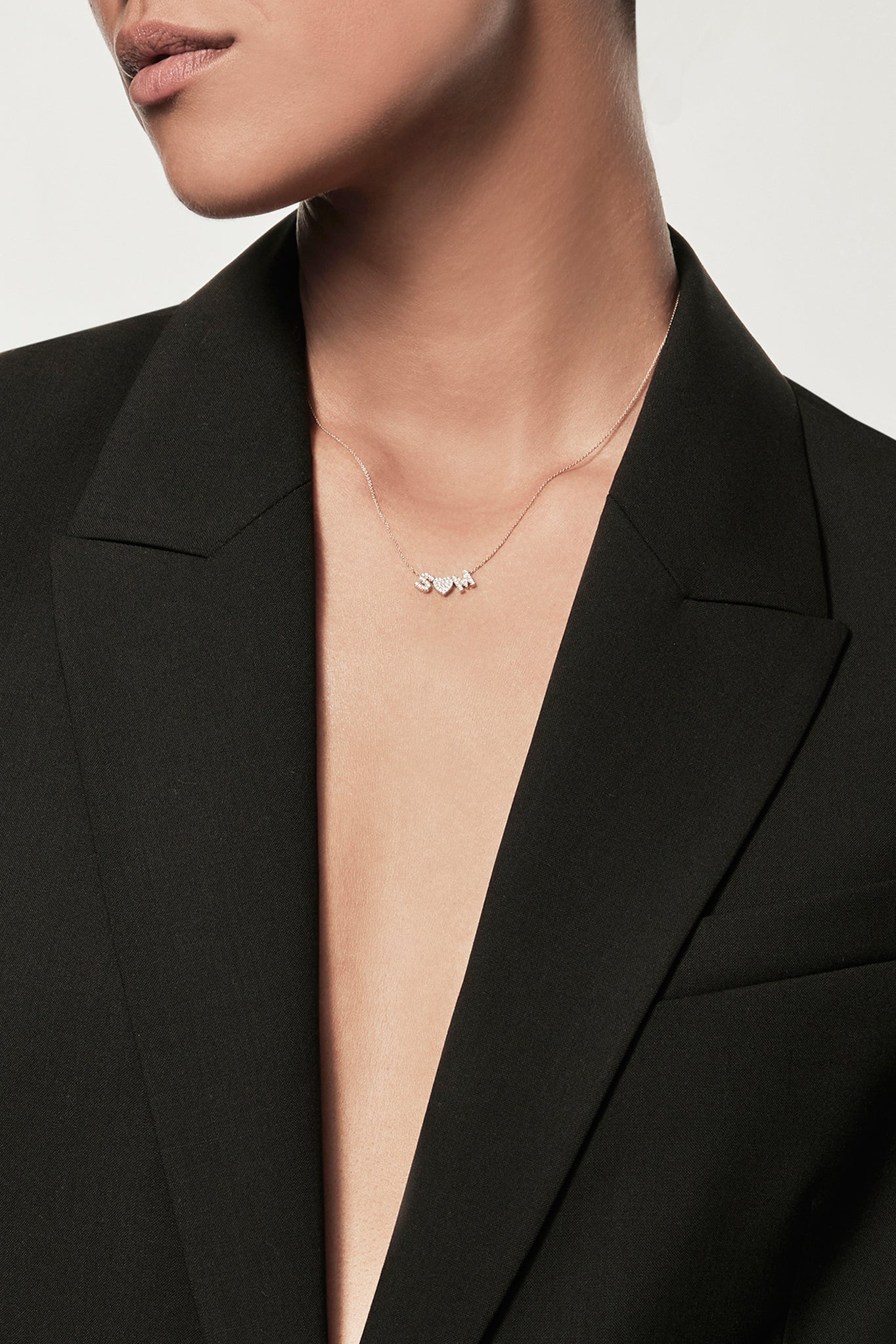 The Two Letters and Heart Necklace in Diamonds and 18k Gold showcases a delicate 18k solid gold chain adorned with three charm pendants: the letters "S" and "M" embellished with small, shimmering diamonds, and a central heart charm adorned with matching diamonds. This elegant piece boasts a minimalist chain without other decorations.