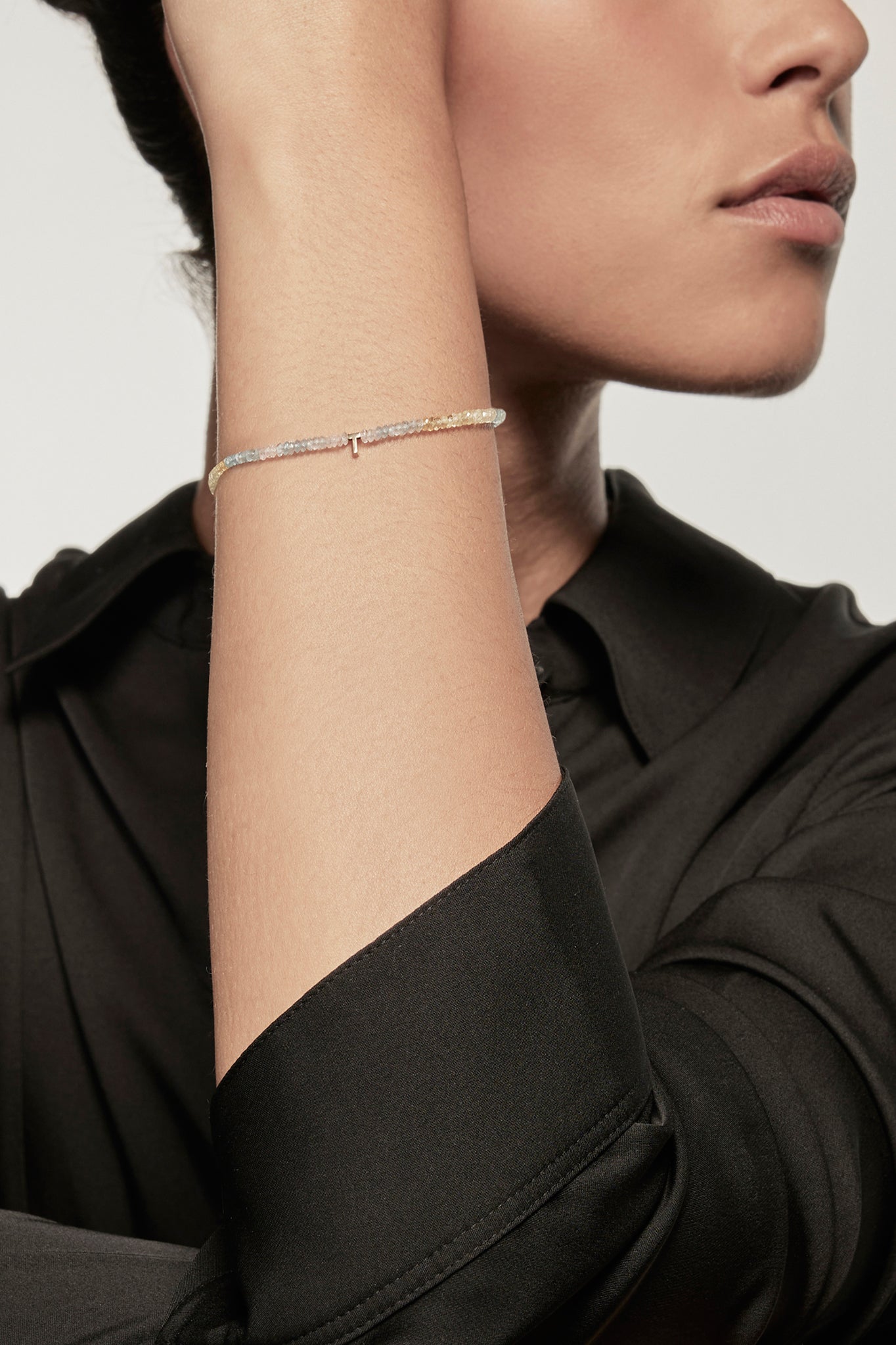 A faceted aquamarine petite bracelet with a gold heart charm is worn on the left wrist of a model dressed in a black silk shirt. The bracelet's deep red garnets contrast with the shirt's dark fabric, and the small gold heart adds a delicate accent.