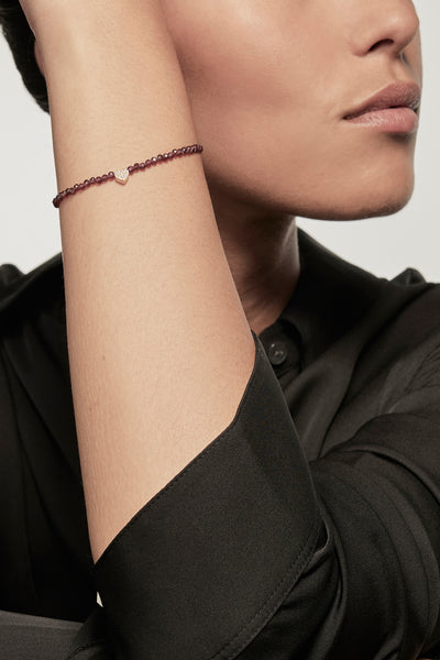 A faceted garnet petite bracelet with a gold heart charm is worn on the left wrist of a model dressed in a black silk shirt. The bracelet's deep red garnets contrast with the shirt's dark fabric, and the small gold heart adds a delicate accent.