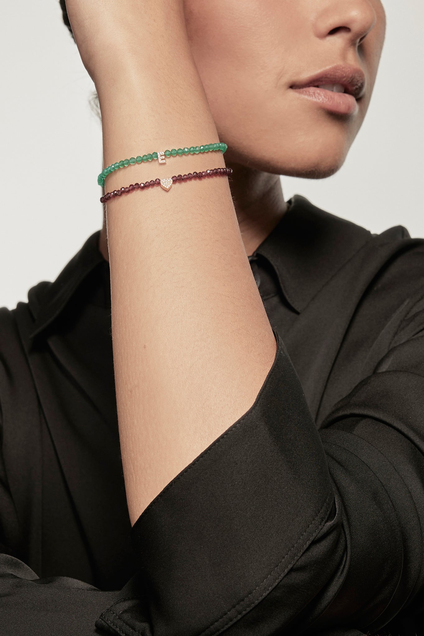 A faceted green onyx petite bracelet with a gold heart charm is worn on the left wrist of a model dressed in a black silk shirt. The bracelet's deep red garnets contrast with the shirt's dark fabric, and the small gold heart adds a delicate accent.