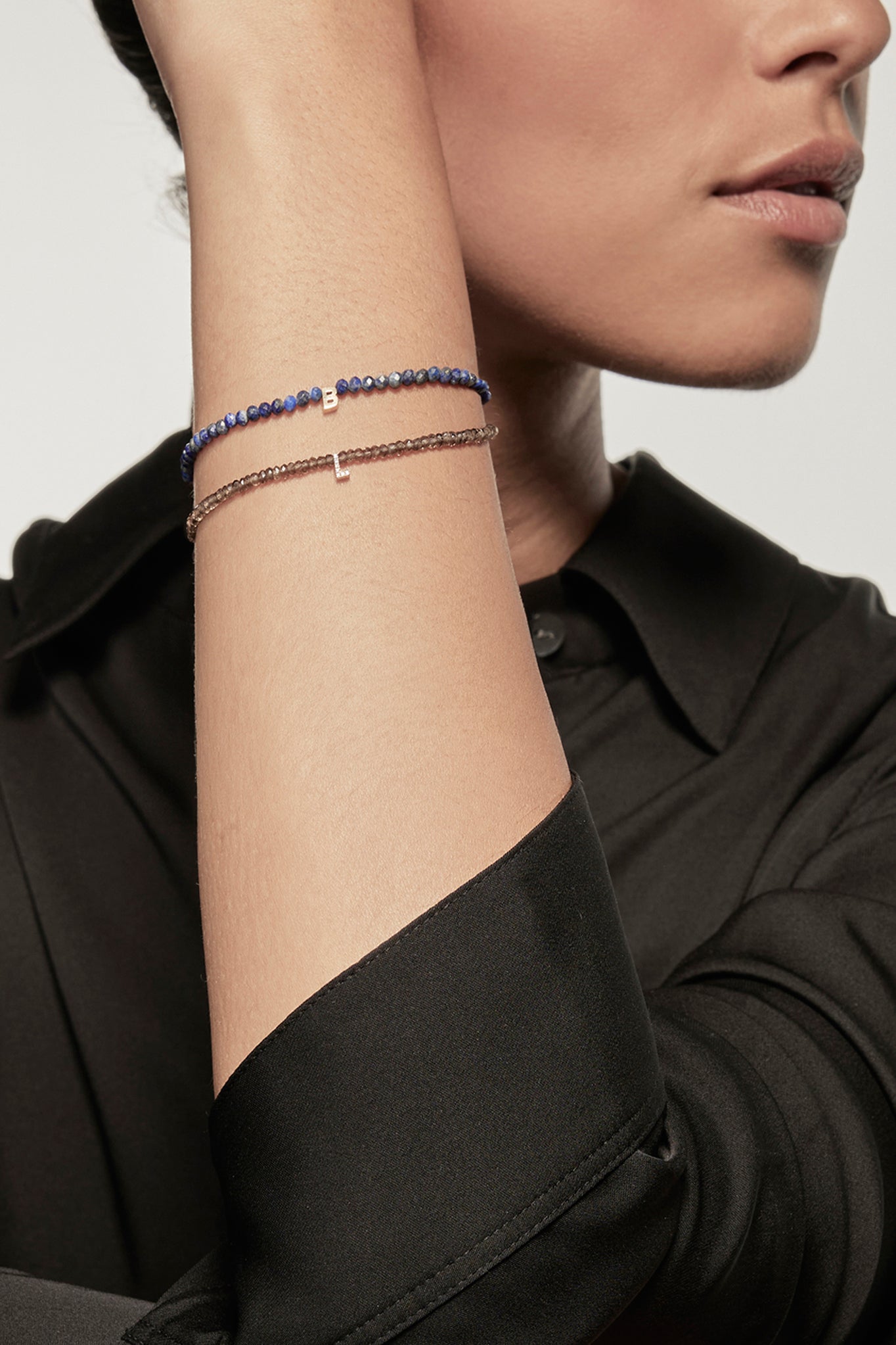 A faceted lapis petite bracelet with a gold heart charm is worn on the left wrist of a model dressed in a black silk shirt. The bracelet's deep red garnets contrast with the shirt's dark fabric, and the small gold heart adds a delicate accent.