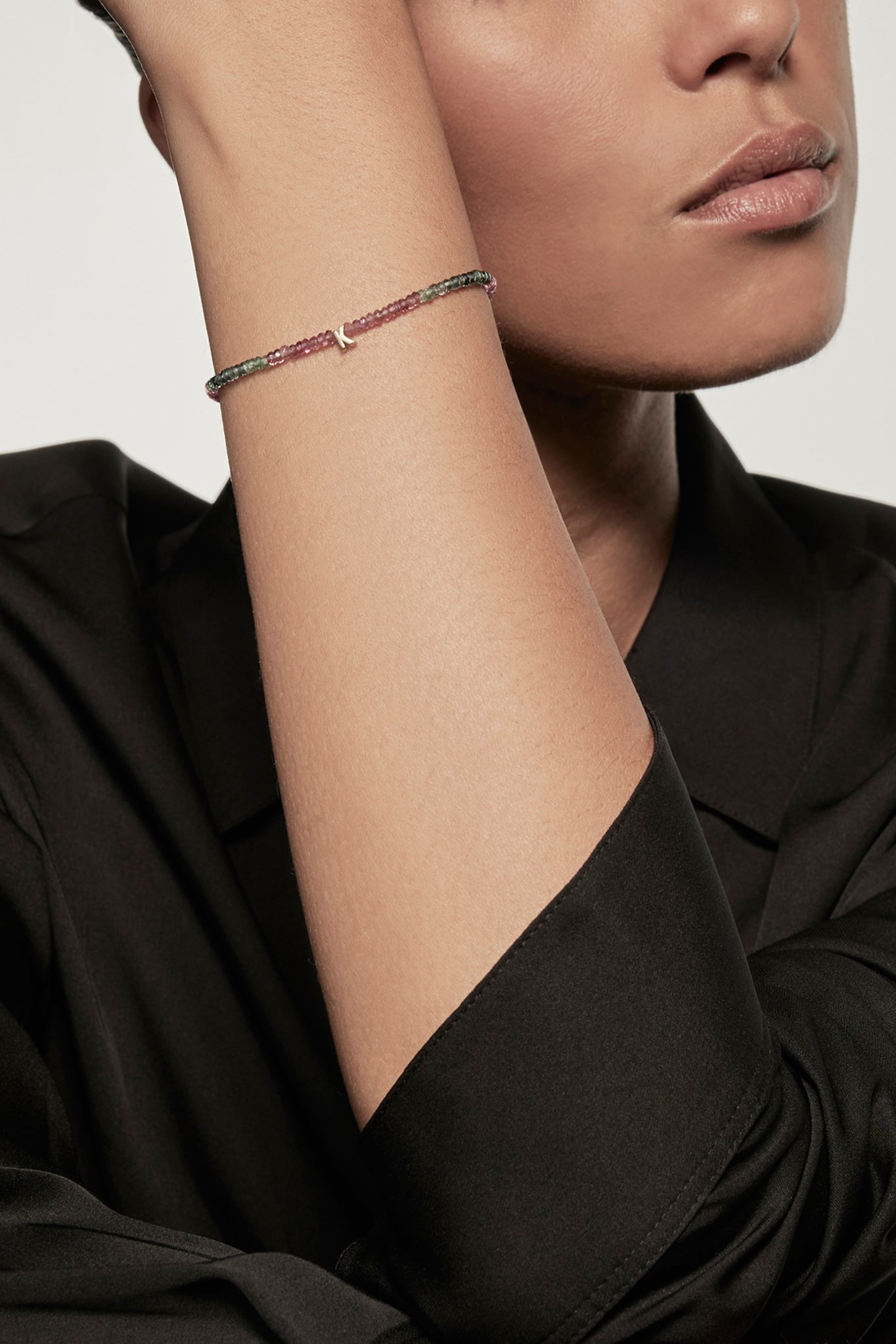 A faceted tourmaline petite bracelet with a gold heart charm is worn on the left wrist of a model dressed in a black silk shirt. The bracelet's deep red garnets contrast with the shirt's dark fabric, and the small gold heart adds a delicate accent.