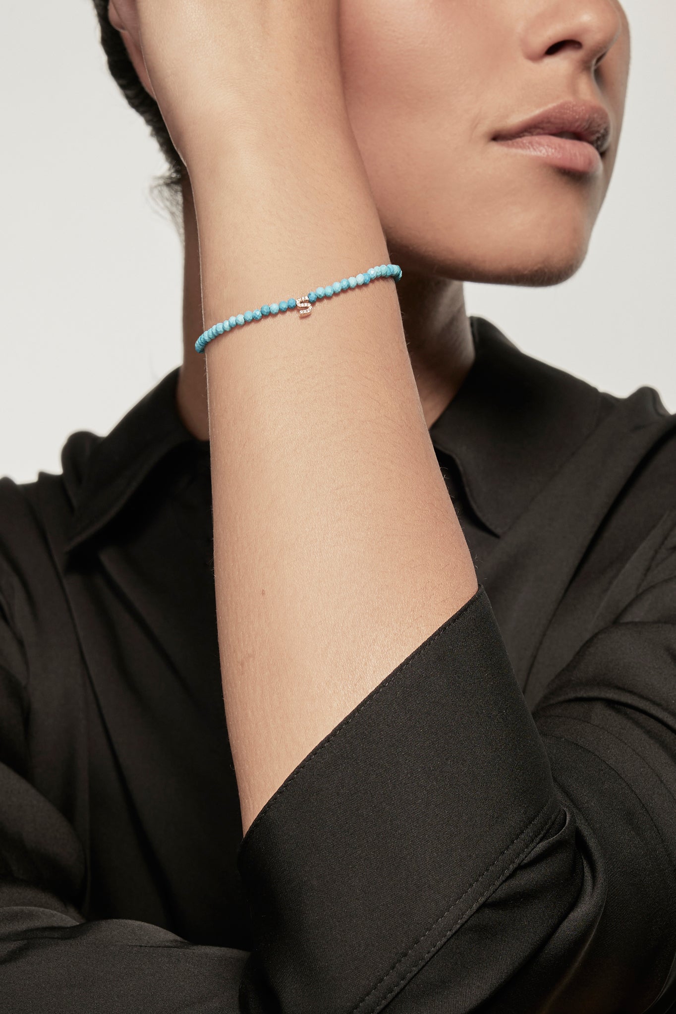 A faceted turquoise petite bracelet with a gold heart charm is worn on the left wrist of a model dressed in a black silk shirt. The bracelet's deep red garnets contrast with the shirt's dark fabric, and the small gold heart adds a delicate accent.
