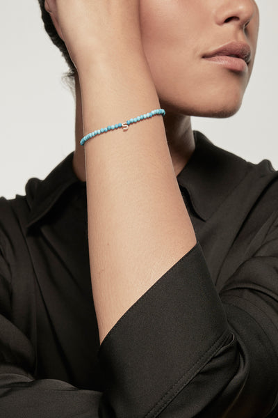 A faceted turquoise petite bracelet with a gold heart charm is worn on the left wrist of a model dressed in a black silk shirt. The bracelet's deep red garnets contrast with the shirt's dark fabric, and the small gold heart adds a delicate accent.
