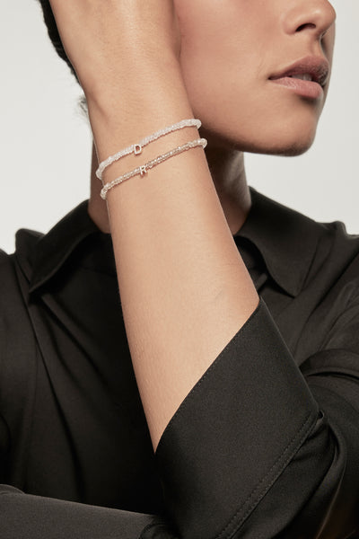 A faceted rainbow moonstone petite bracelet with a diamond heart charm is worn on the left wrist of a model dressed in a black silk shirt. The bracelet's deep red garnets contrast with the shirt's dark fabric, and the small gold heart adds a delicate accent.