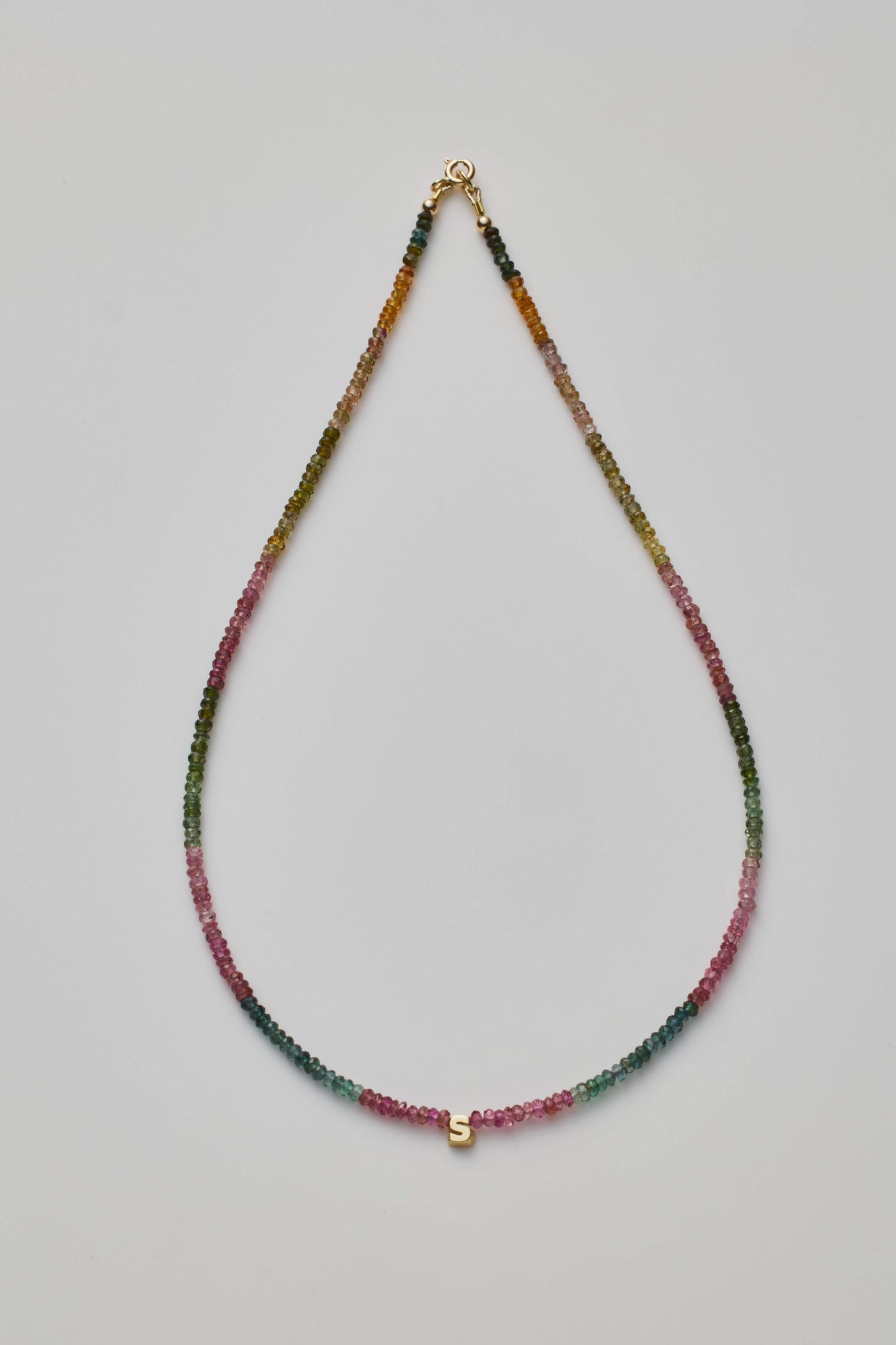 The One Letter Necklace in Tourmaline and 14k Gold is a delicate beaded necklace featuring a gradient pattern of small, multi-colored tourmaline gemstones transitioning from greens to pinks. At its center, the necklace showcases a small 14k gold 'S' charm, set against a plain white background.